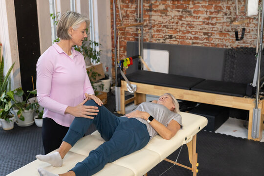 Senior female therapist supporting senior female patient on padded table in rehab studio