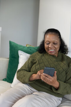 Senior African American woman using phone on sofa with green and white pillows, in green sweater