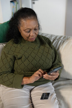 Senior African American woman sitting at home in olive sweater tapping phone, holding card