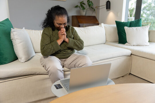 Senior African American woman sitting on sofa in living room in olive sweater using silver laptop