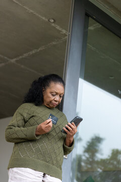 Senior African American woman standing in door by glass using phone holding card