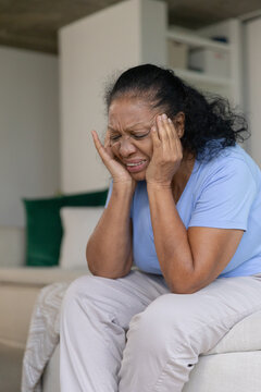 Senior African American woman sitting on sofa at home holding temples, wearing blue tee with pillow