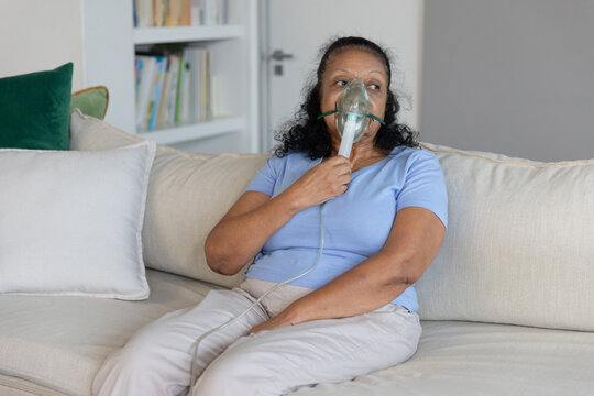 Senior African American woman holding oxygen mask tubing while sitting on sofa in living room