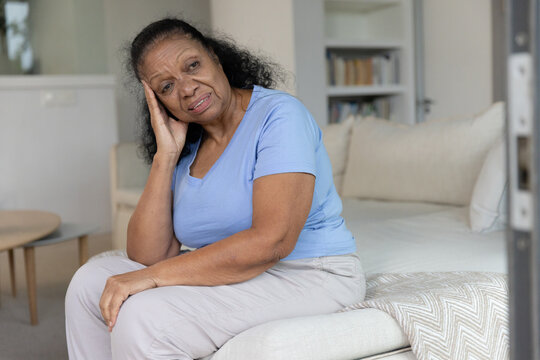 Senior African American woman sitting on bed holding head, wearing blue top and beige pants