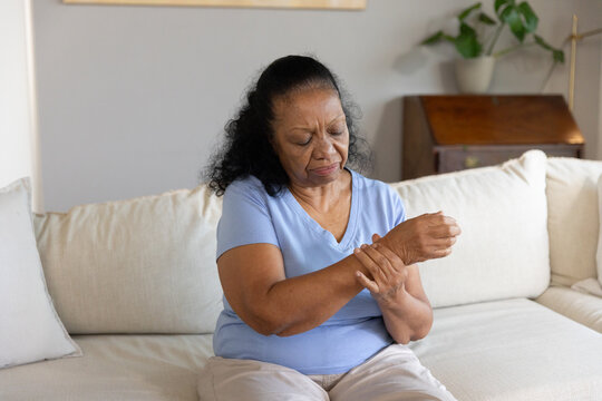 Senior African American woman sitting on cream sofa in blue top massaging right wrist near plant