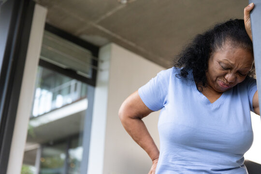 Senior African American woman in light blue V neck clutching back, leaning on post, copy space