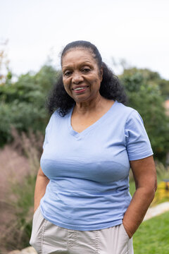Senior African American woman standing in garden in light blue shirt, beige pants, hands in pockets