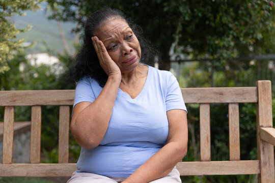 Senior African woman sitting on wooden bench in garden, holding cheek and wearing V-neck shirt