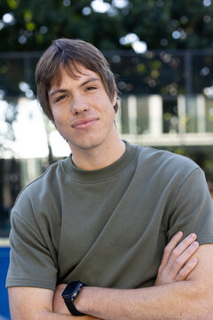 Mid20s man posing arms crossed wearing olive tee and black smartwatch by glass facade showing trees