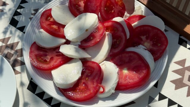 sliced red tomatoes and white mozzarella cheese arranged on a white ceramic plate, placed on a patterned fabric tablecloth