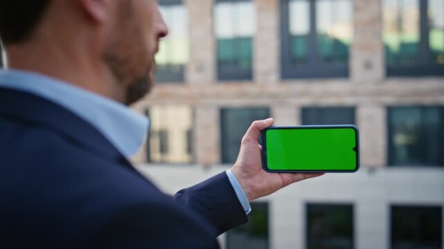 Businessman holding smartphone showing green screen on rooftop balcony in modern city, arm reaching toward camera during presentation gesture at daylight outdoors