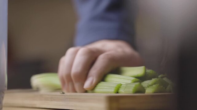 Hand slicing celery stalks into even rounds on wooden cutting board with close hand and knife detail in soft natural side light, shallow depth of field during kitchen prep
