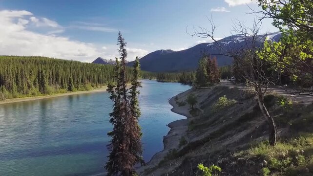 River bend flowing past gravel and eroded banks toward snow-capped mountain ridge beneath partly cloudy sky with sunlit forest and tranquil current