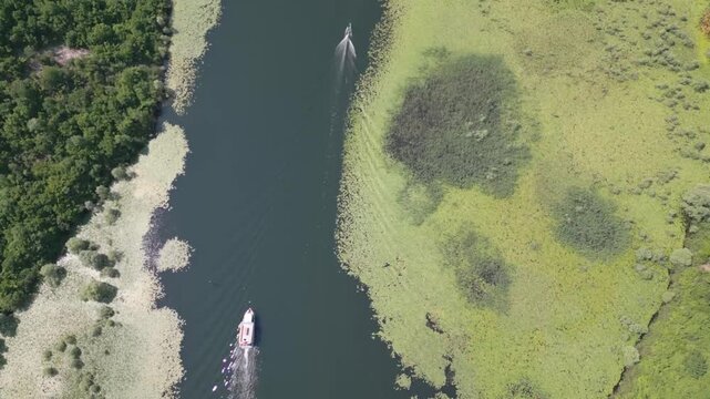 Aerial top-down view of small motorboat moving through narrow meandering river with trailing wake and sun glint