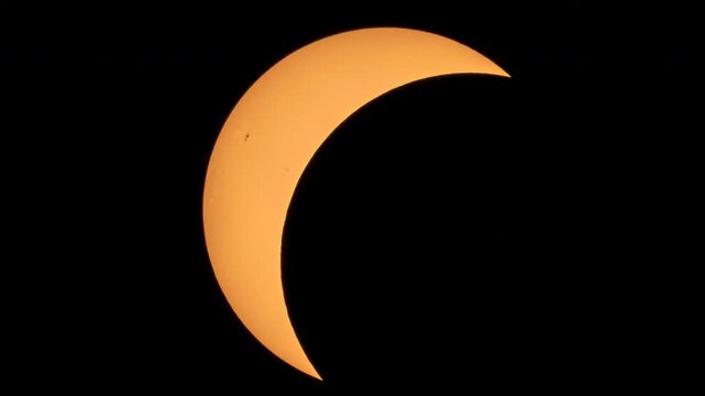 A dramatic partial solar eclipse against a dark sky, with the moon covering most of the vibrant orange sun, revealing a crescent and sunspots