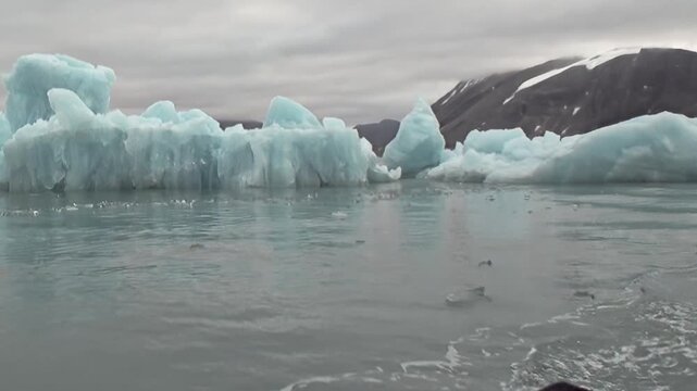 A blue iceberg floats on cold water beneath rugged mountain slopes. Additional visible detail includes snow and wide framing, with natural light defining the surrounding forms.