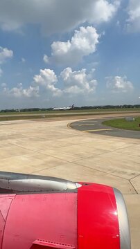 Airplane wing and red engine visible from the window as the aircraft prepares for takeoff, showcasing the runway and scattered clouds under blue sky. Slow motion vertical footage, 4K, copyspace