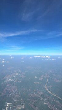 Aerial view of green landscape with scattered clouds and blue sky captured from an airplane window during daytime flight. Slow motion vertical footage, 4K, copyspace