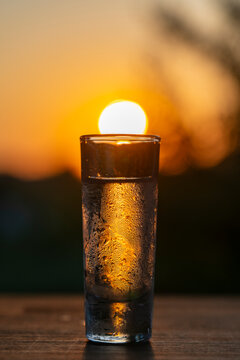One frosted shot glass with vodka against sunset sun, closeup. Ice cold tequila or vodka shot covered in frost and condensation on wooden table over beautiful golden sunset sky with sun in background