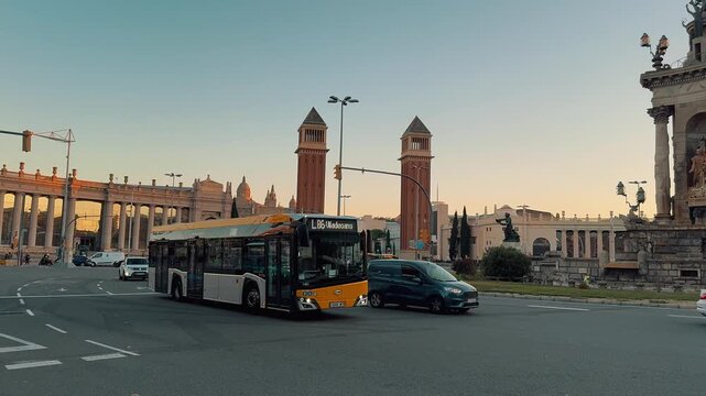 Bustling city traffic and pedestrians cross around iconic Plaza Espana at sunset, featuring the historic Venetian Towers and magnificent Palau Nacional atop Montjuic hill in Barcelona, Catalonia,