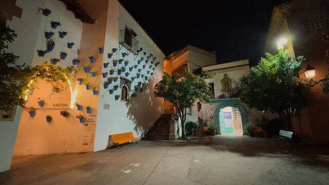 Explore the illuminated streets of Poble Espanyol at night in Barcelona, showcasing traditional Spanish architecture, fortified medieval gate, decorative hanging umbrellas, historic facades,