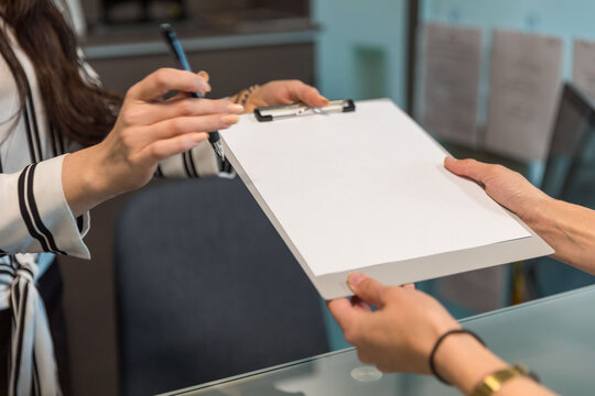 Two people exchanging a clipboard over a glass desk in an office.