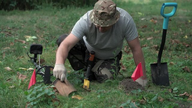 Male treasure hunter in camouflage pants and gray shirt uses a small digging tool to uncover buried items in a grassy area marked with flags, with a metal detector and shovel nearby