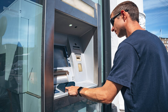 A man in casual clothing uses an automated teller machine on a sunny street to withdraw cash or check his balance, representing personal finance management and modern banking convenience.