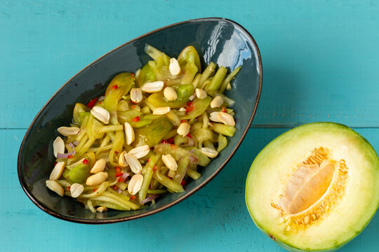 Thai Som Tam-style melon salad on an oval platter, sprinkled with peanuts, next to an unripe Charentais half on blue boards.