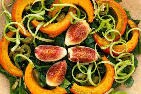 A close-up top view of an autumnal salad featuring baked pumpkin, figs, and curled puntarelle stalks atop blanched New Zealand spinach leaves.