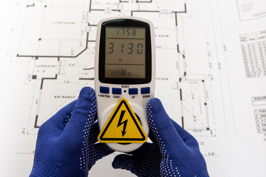 A worker holds a portable voltmeter against the backdrop of a paper floor plan of an apartment; the voltmeter's socket is covered by a high-voltage warning sign.