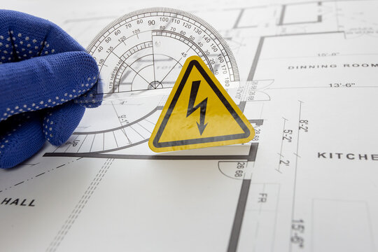 A high-voltage warning sign is being held against a paper drawing by an engineer using a protractor ruler.