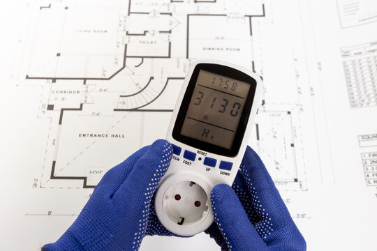 An electrician holds a portable current meter over a paper blueprint of a house.