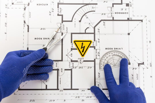 A close-up, high-angle shot shows an electrician holding a caliper and a protractor with gloved hands over a paper floor plan of an apartment, upon which a high-voltage warning sign is placed.