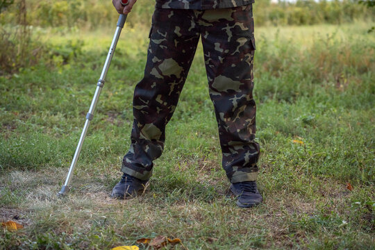 A confident military veteran, dressed in a camouflage outfit, stands in a lush green field. He uses a cane for support while demonstrating resilience and strength in the bright sunlight