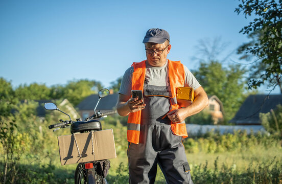 A delivery worker wearing an orange vest stands by his bicycle in a sunny rural setting. He is focused on his phone, likely confirming delivery details while enjoying the greenery