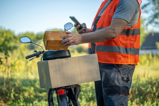 A delivery person in a bright orange vest balances two packages on a bicycle. Surrounded by green fields, they check their phone, reflecting a busy workday in nature
