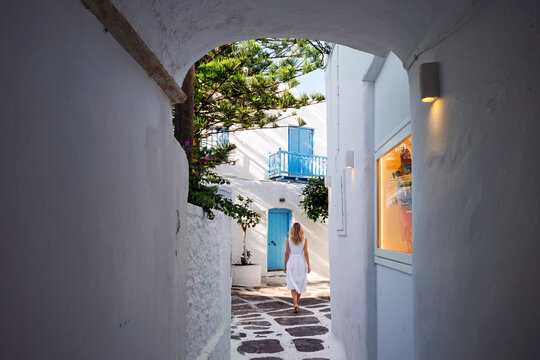 A young woman walks down a narrow whitewashed arched alleyway with colorful doorways and shutters in the old town center of Mykonos, Greece.