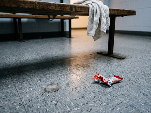 Discarded trash and litter on the floor of a messy locker room. Crumpled snack wrapper and plastic cap under a wooden bench with a towel. Unsanitary gym changing room facility