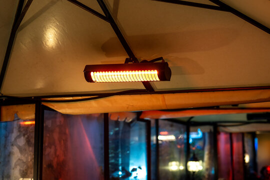 Suspended infrared heater mounted on umbrella in outdoor cafe terrace during autumn-winter season, electric patio heater hanging above ceiling at open-air restaurant lounge area spreading warm air.