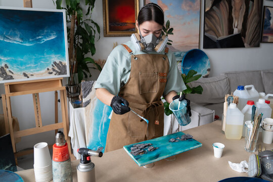 Female artist holds plastic cup filled with blue epoxy resin and carefully drips liquid onto painting with wooden spatula, controlling placement to create desired pattern in home loft workshop.