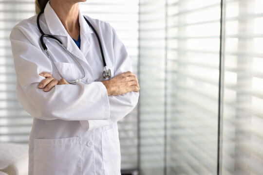 Confident female doctor in white coat and stethoscope standing with folded arms next to window in clinic office, cropped closeup, copy space. Reliability in healthcare, professional medical services