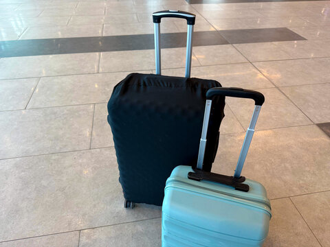 Two suitcases in an airport terminal, one black with a protective cover and one light blue, positioned on a tiled floor with visible reflections and lighting