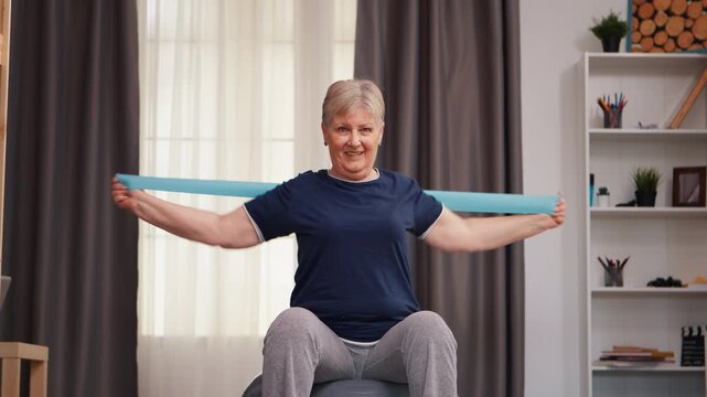 Smiling senior woman exercises with a resistance band while seated on a stability ball at home for gentle strength, balance and healthy aging, while supporting mobility and active aging.