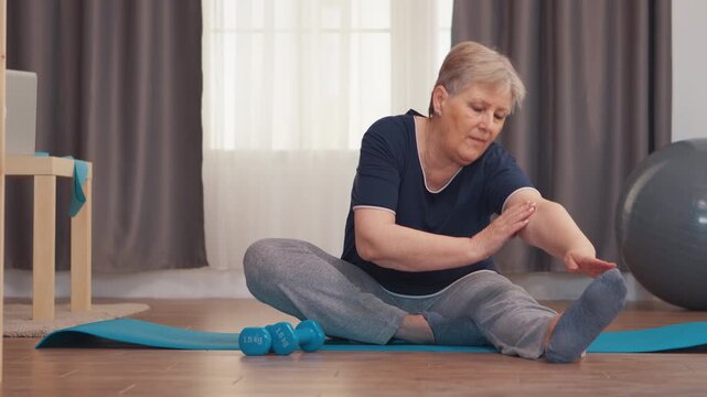 Senior woman reaches toward her foot in a seated leg stretch on a yoga mat, supporting flexibility, mobility and active aging through home exercise.