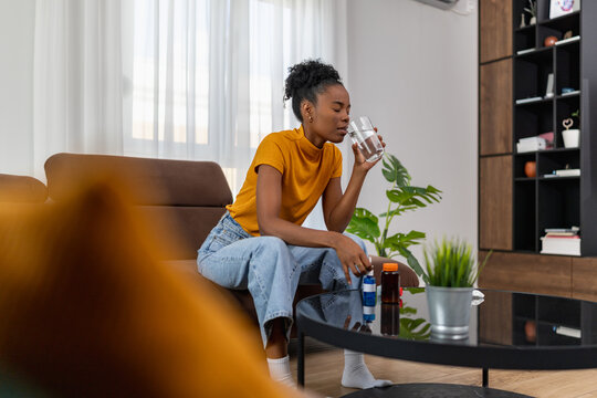 Young woman sitting on couch drinking water while taking medicine at home