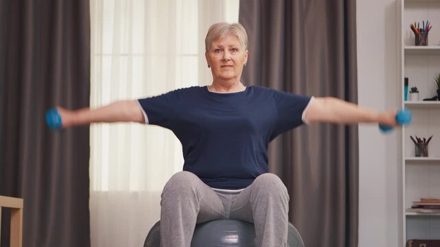 Senior woman exercises with dumbbells while seated on a stability ball at home, using controlled arm movements for strength, balance and active aging to support mobility and balance training.