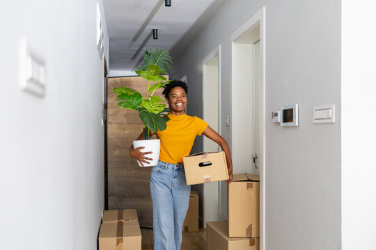 Black woman carrying houseplant and moving boxes while smiling in bright modern hallway