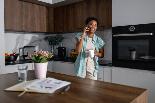 Young woman talking on phone in modern kitchen while holding coffee mug and smiling