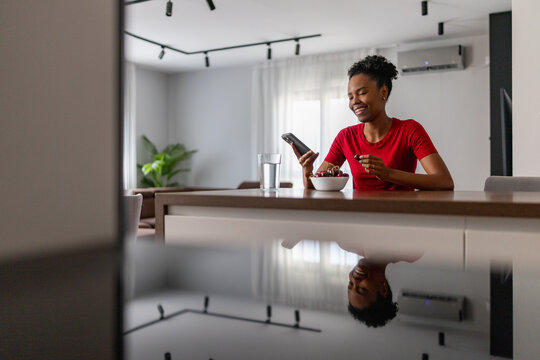 Smiling woman eating cherries while checking smartphone at modern kitchen counter